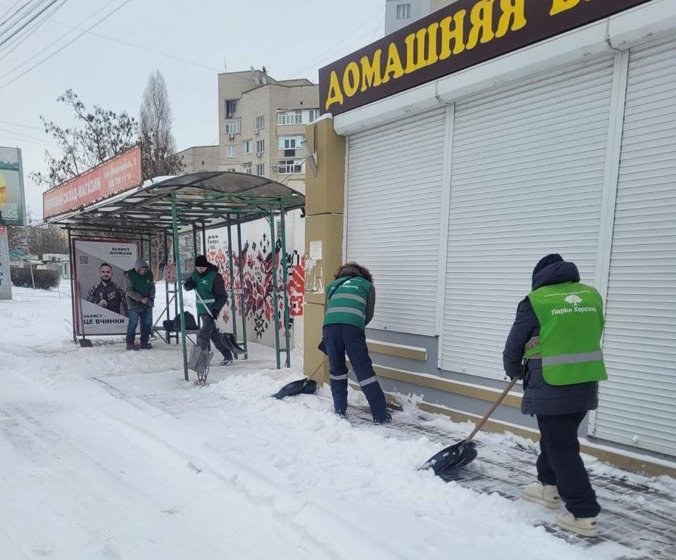 Комунальні працівники у світловідбивальних жилетах розчищають тротуар від снігу біля зупинки громадського транспорту та зачиненого магазину.