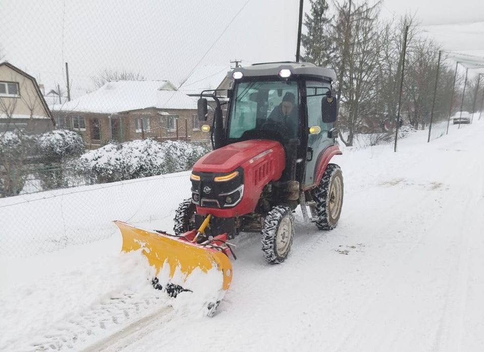 Червоний трактор із жовтим відвалом розчищає дорогу від снігу в приватному секторі під час снігопаду.