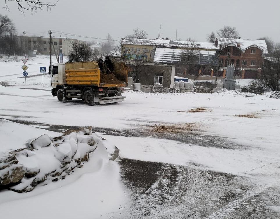 Вантажівка з піщано-сольовою сумішшю рухається засніженою дорогою в населеному пункті. Працівники в кузові розкидають суміш лопатами для боротьби з ожеледицею на фоні приватних будинків.