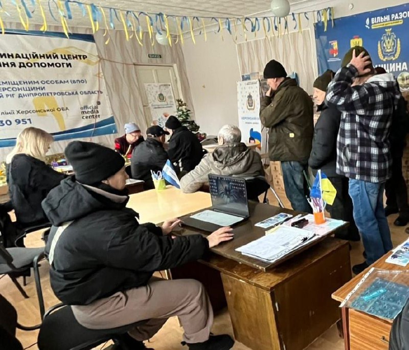 Люди в приміщенні координаційного центру допомоги переселенцям. Працівники сидять за столами з ноутбуками та документами, надаючи консультації відвідувачам. На стінах розміщені інформаційні банери з українською символікою та контактами гарячої лінії.