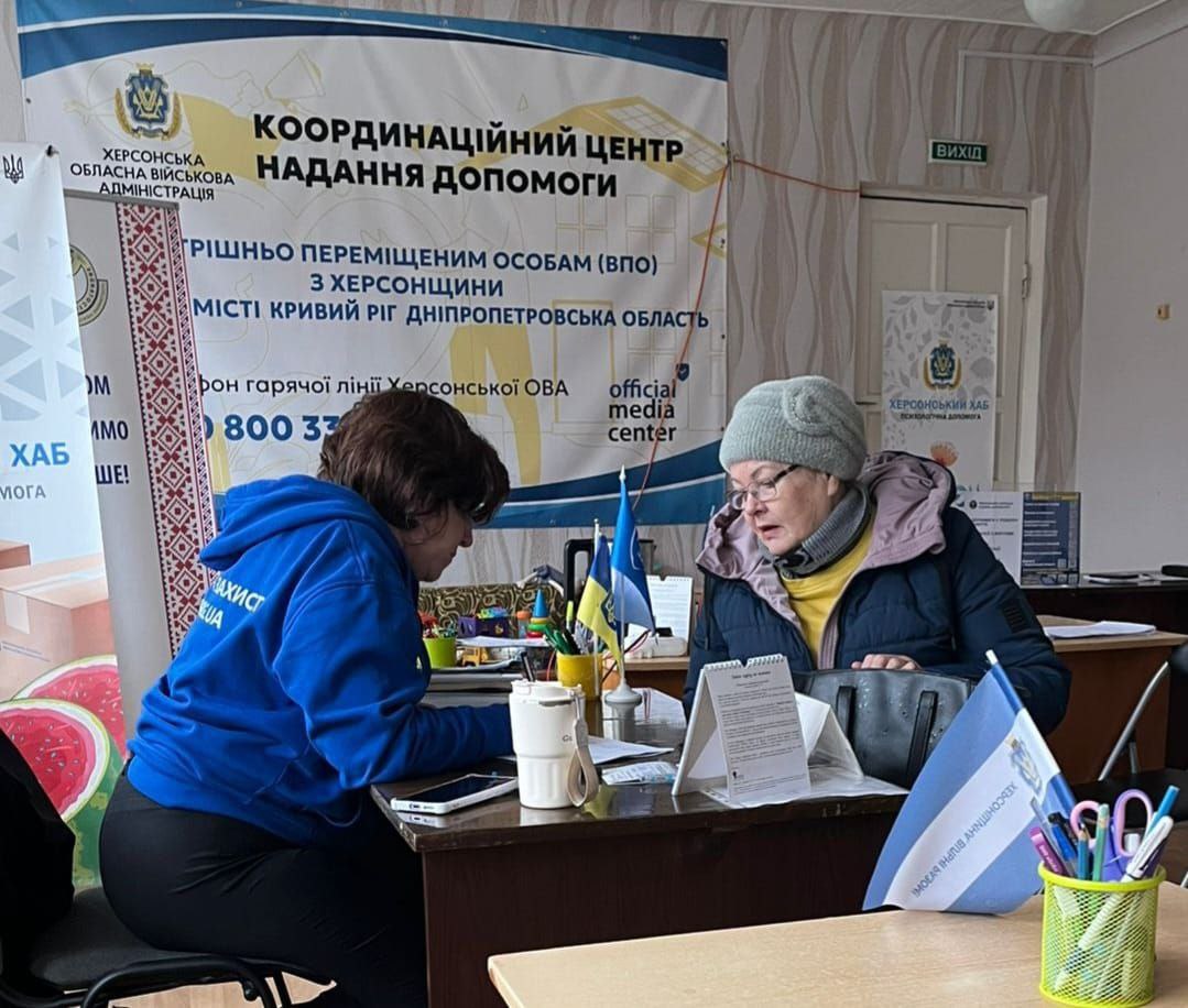 Працівниця координаційного центру допомоги переселенцям з Херсонщини консультує відвідувачку за робочим столом. На фоні розміщено банер із контактами гарячої лінії Херсонської ОВА та інформацією про хаб у Кривому Розі.