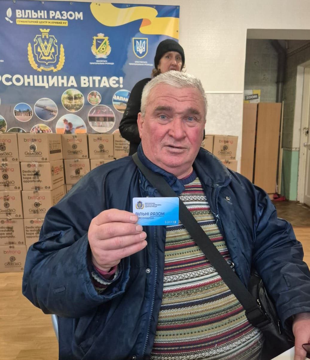 Чоловік у гуманітарному центрі тримає в руках пластикову картку «Вільні разом». На фоні видно коробки з допомогою та банер із гербом Херсонської області та написом «Херсонщина вітає!».