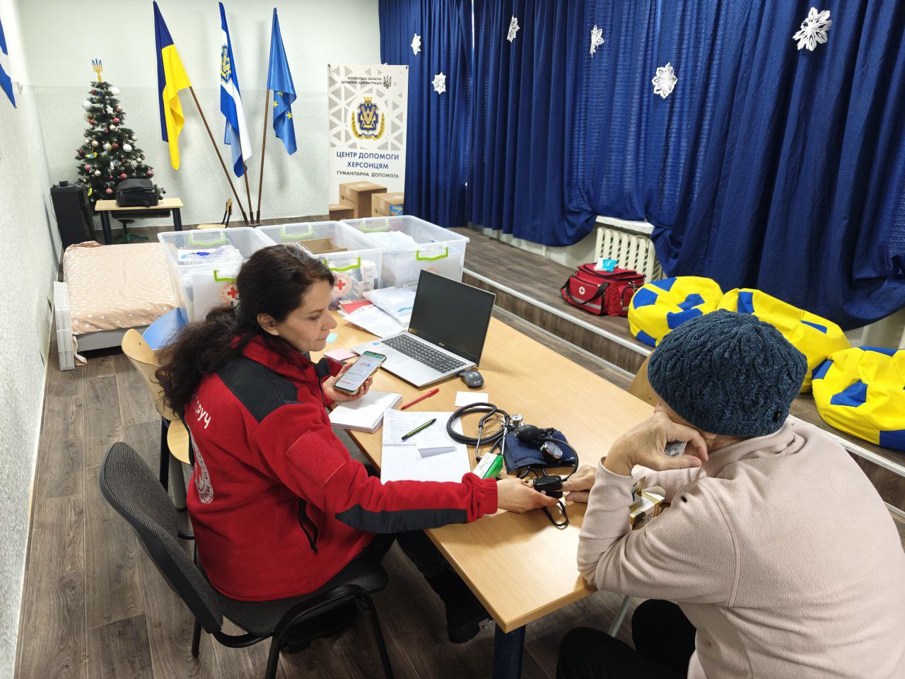 Працівниця гуманітарного центру в червоному одязі вимірює пульс або рівень кисню жінці у бежевій кофті та синій в'язаній шапці за допомогою портативного пристрою. На столі лежать тонометр, ноутбук та документи. У приміщенні з синіми шторами та святковим декором також видно контейнери з допомогою, прапори України та ЄС і плакат «Центр допомоги херсонцям».
