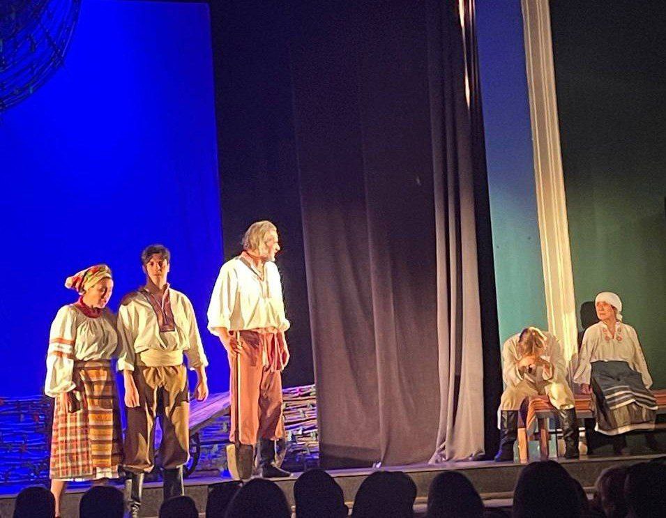 Фото театральної вистави на сцені. П'ятеро акторів у традиційному українському народному вбранні виконують драматичну сцену. На задньому плані — синє освітлення та декорації сільського побуту.