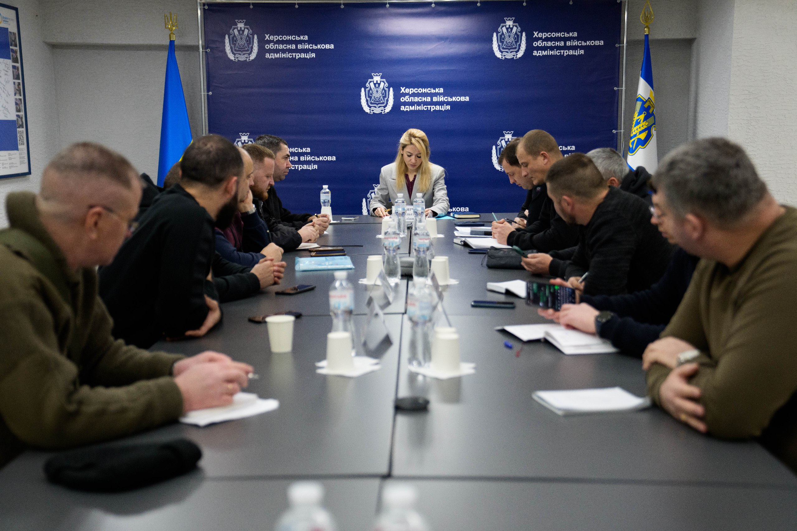 Робоча нарада в Херсонській обласній військовій адміністрації. Керівниця та учасники засідання сидять за довгим столом у конференц-залі на фоні брендованого синього банера з гербом області та офіційною назвою установи.