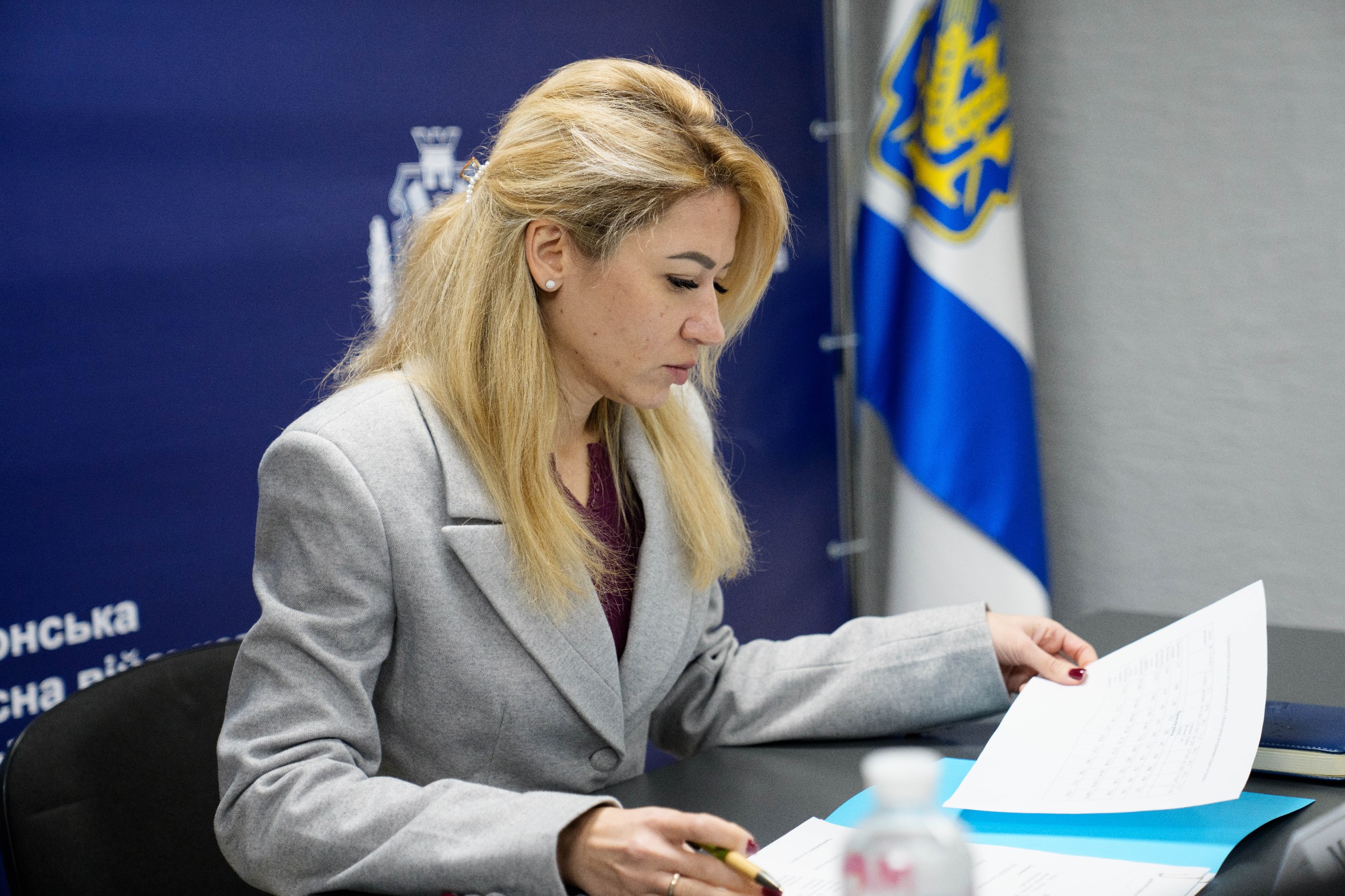 Жінка у світлому піджаку працює з документами за столом у конференц-залі. На фоні розміщено синій банер із гербом та офіційною назвою Херсонської обласної військової адміністрації.