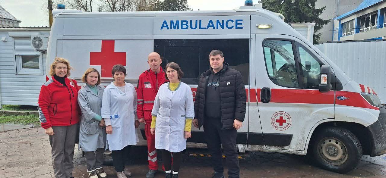 Група медичних працівників та волонтерів стоїть перед автомобілем швидкої допомоги з символікою Червоного Хреста. На білому фоні машини розміщено великий червоний хрест та напис «AMBULANCE».