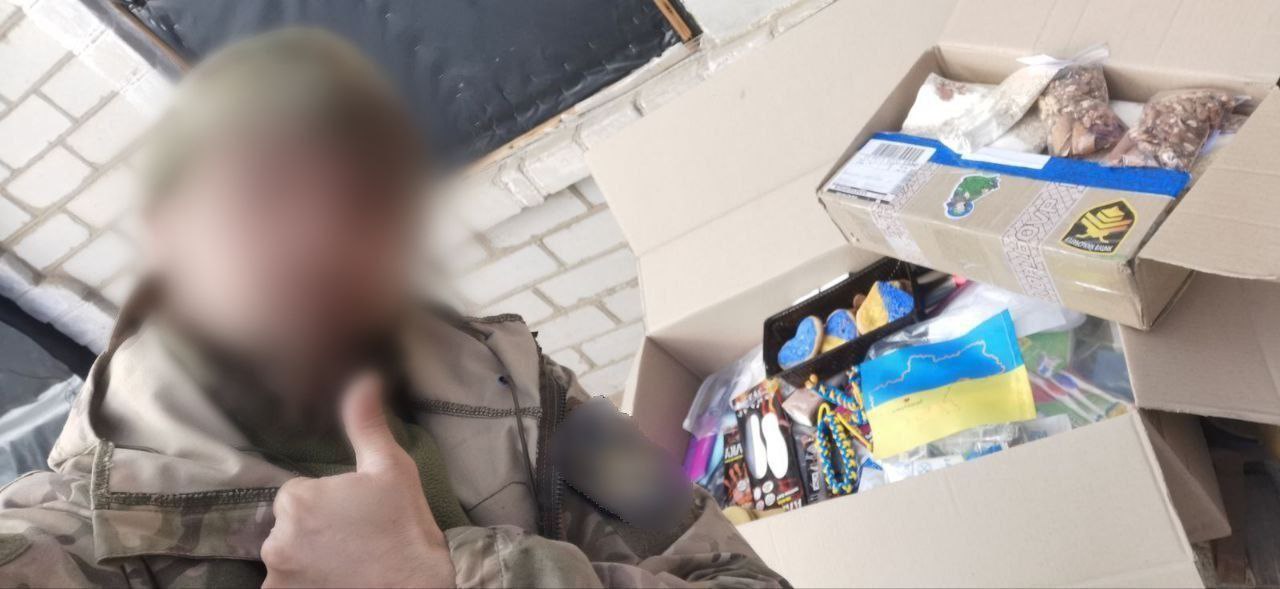 Військовий у камуфляжі з розмитим обличчям показує жест «клас» поруч із відкритою коробкою волонтерської допомоги. У коробці видно продукти харчування (горіхи, сухофрукти) та патріотичну символіку: синьо-жовтий прапорець і в’язані серця.