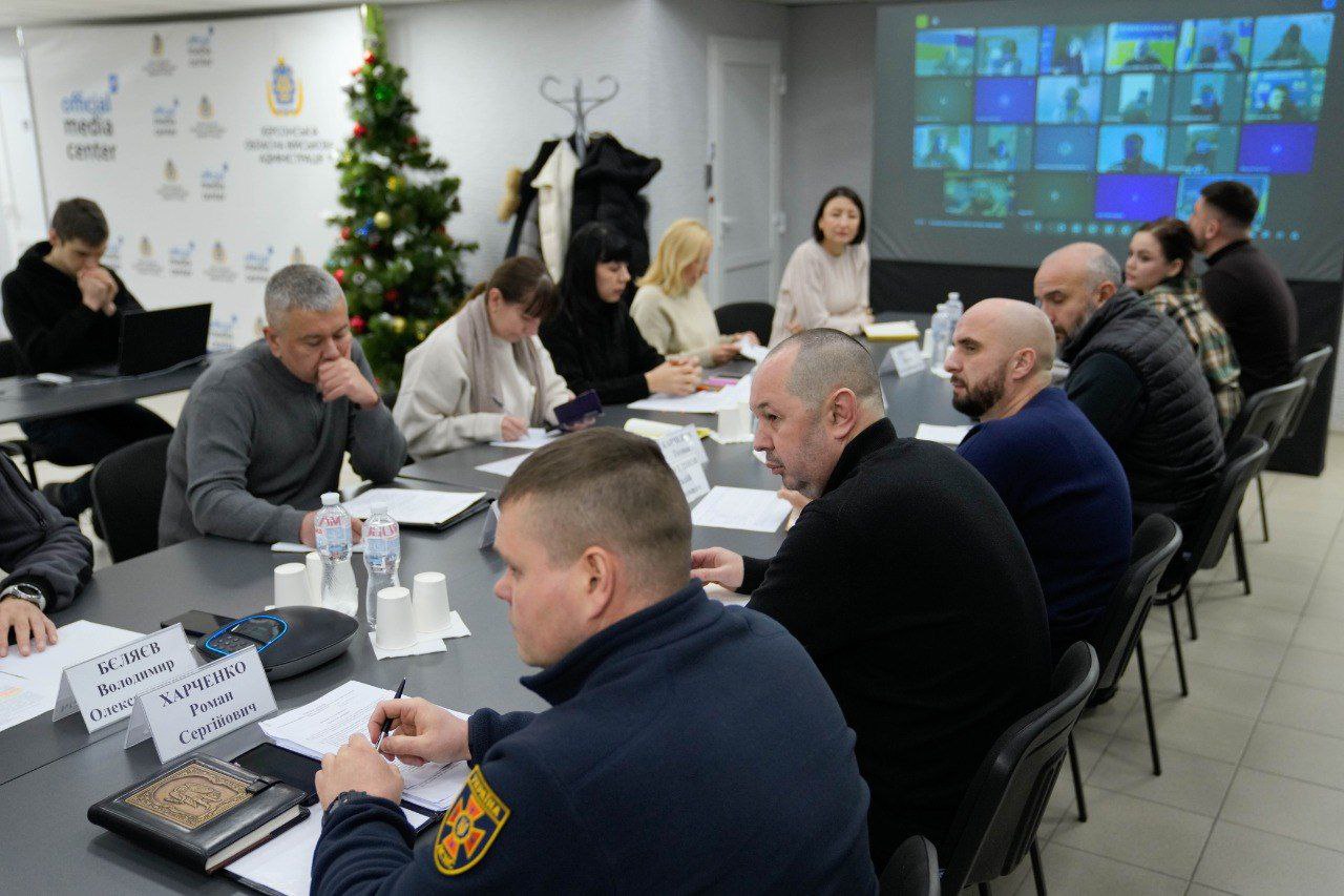 Група людей у цивільному та офіційному одязі сидить за довгим столом під час наради. Учасники роблять записи та переглядають документи. На задньому плані видно прикрашену новорічну ялинку та великий екран, на якому транслюється відеоконференція з багатьма учасниками.