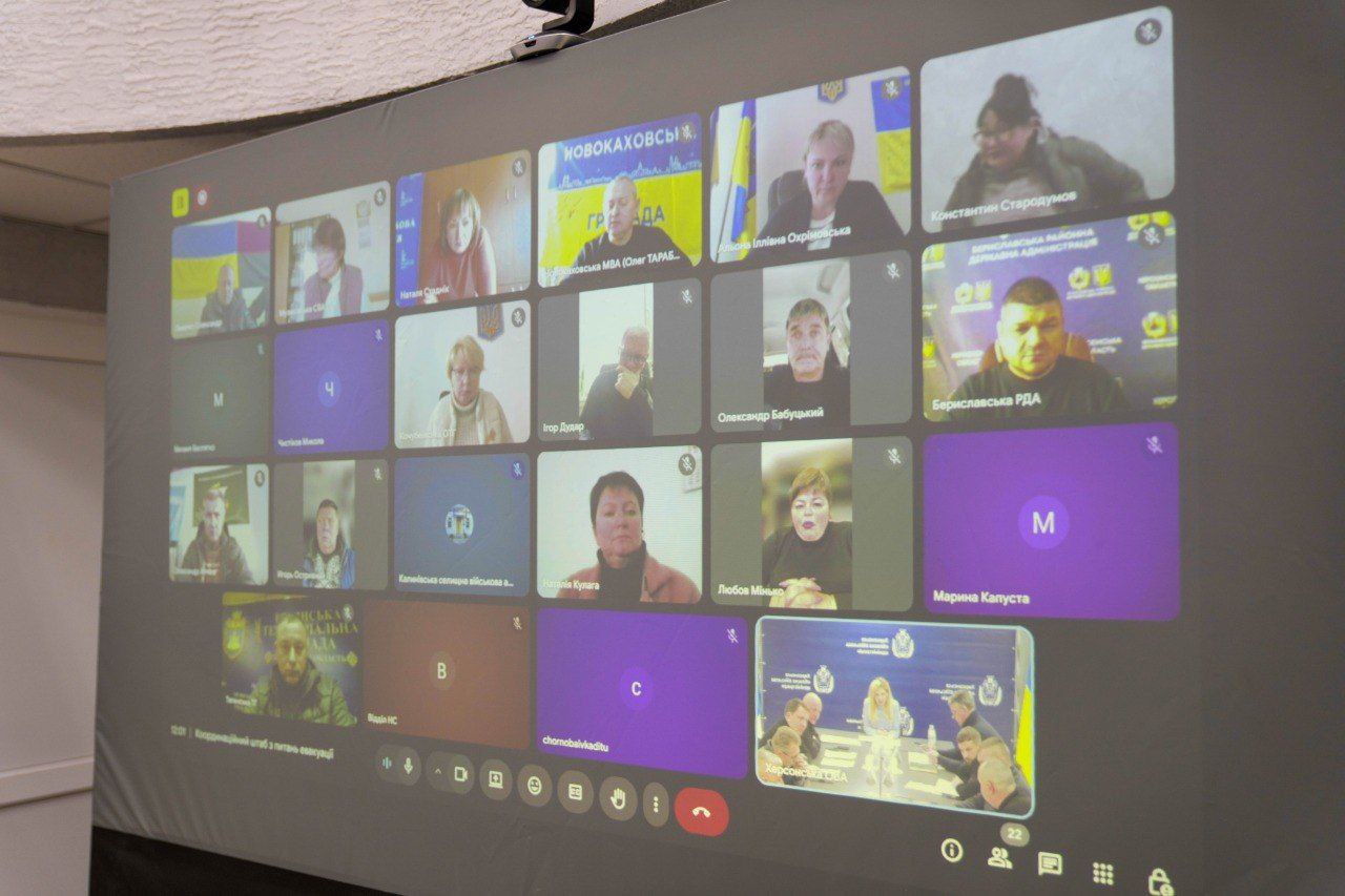 Великий проєкційний екран, на якому відображено вікно відеоконференції з багатьма учасниками. Більшість вікон показують людей на фоні державних прапорів або офіційних банерів місцевих адміністрацій, зокрема Бериславської РДА та Херсонської ОВА. Деякі вікна містять лише іконки з літерами або назвами відомств.