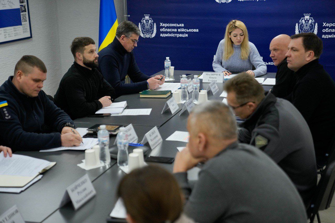 Група офіційних осіб у цивільному та військовому одязі проводить робочу нараду за столом у кабінеті. Учасники переглядають документи, перед ними встановлені іменні таблички та пляшки з водою. На фоні розміщено прапор України та синій банер із гербом і написом «Херсонська обласна військова адміністрація».