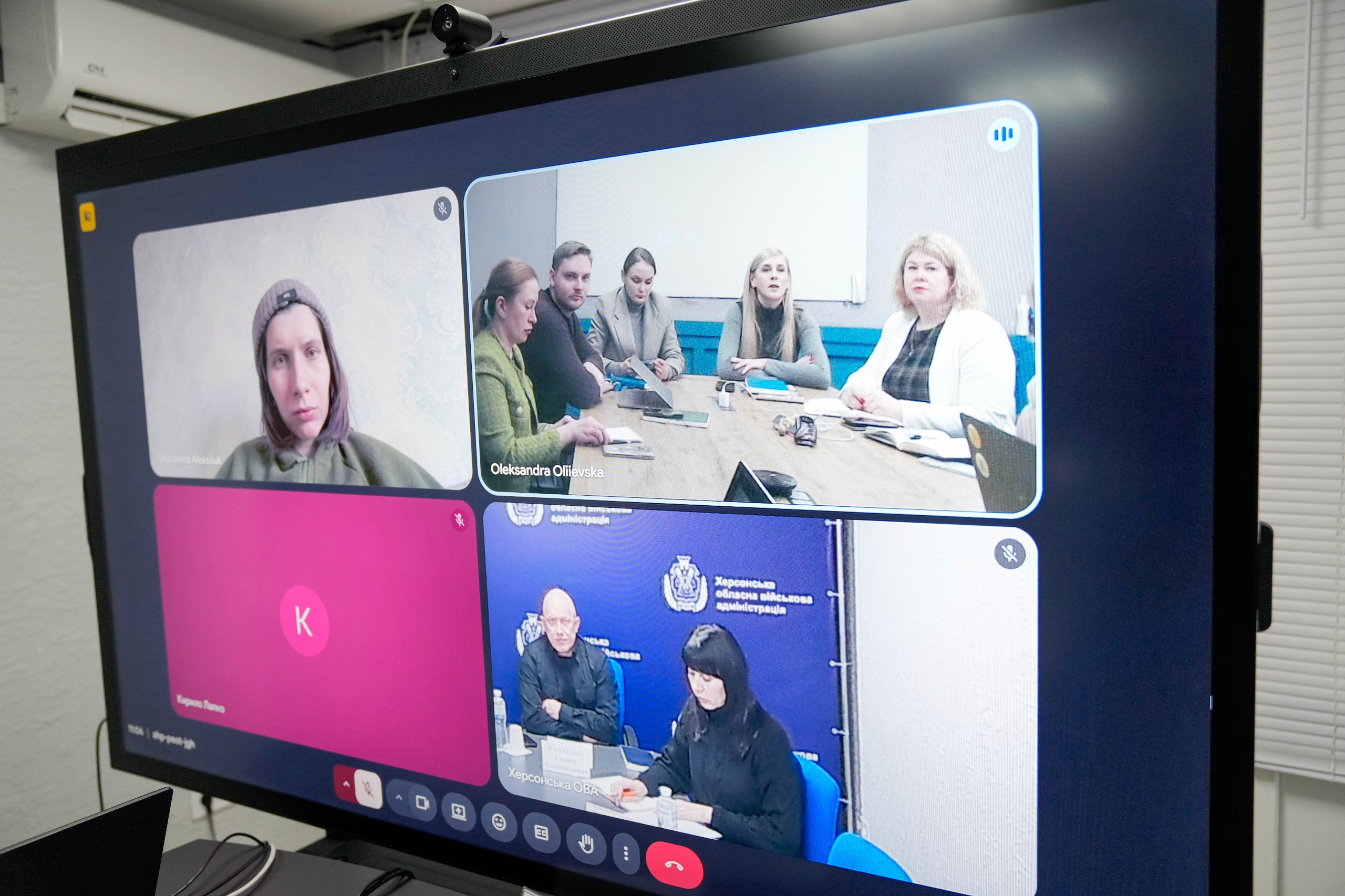 Великий монітор, на якому відображено вікно відеоконференції з чотирма зонами. В одній із них видно групу людей за столом у кабінеті, в іншій — двох учасників на фоні банера Херсонської ОВА, ще одна зона показує учасницю великим планом, а четверта містить лише іконку з літерою. На моніторі встановлено веб-камеру.