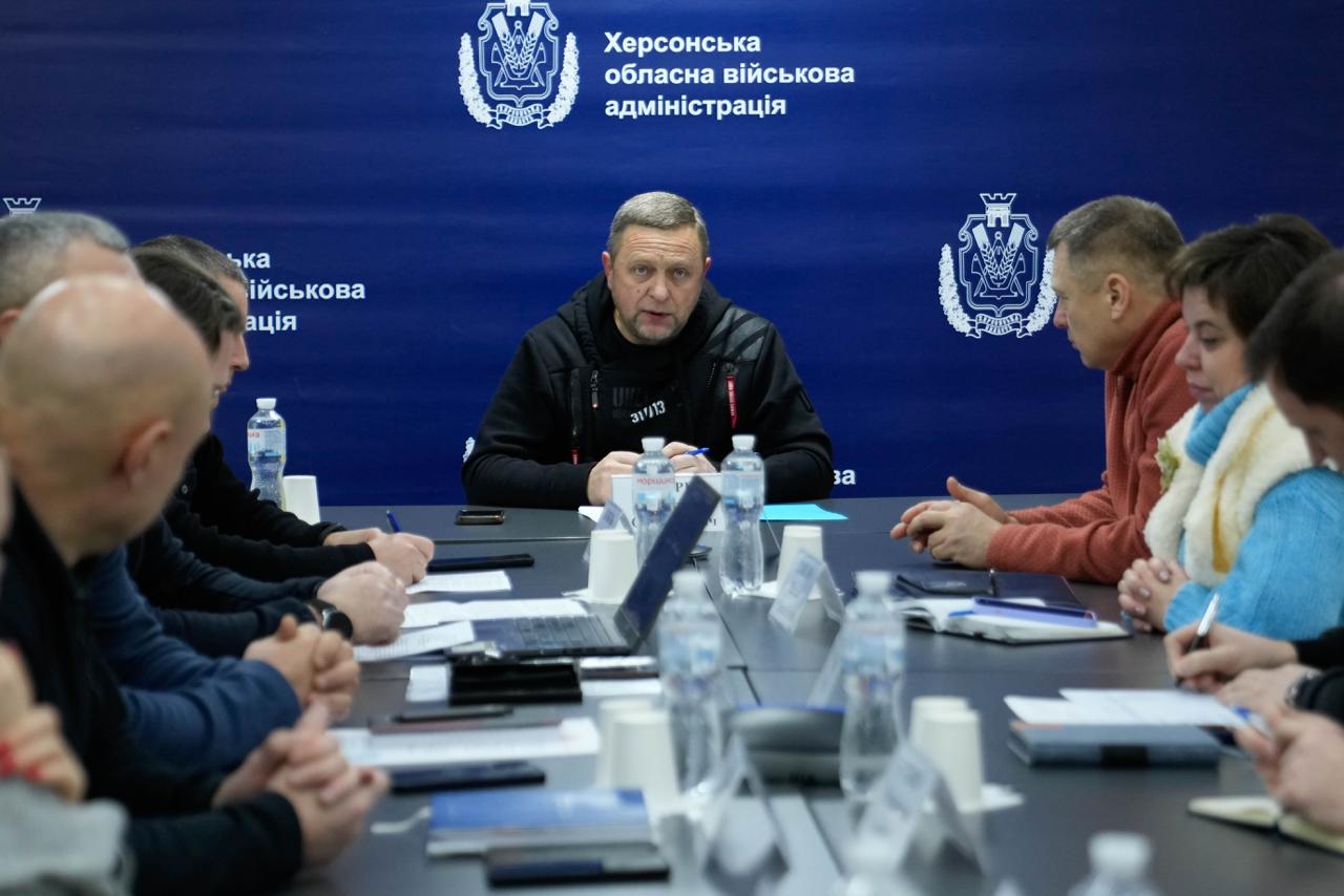 Робоча нарада в приміщенні Херсонської обласної військової адміністрації. Чоловік у чорній спортивній куртці сидить на чолі столу на фоні синього брендованого банера з гербом. За довгим столом по обидва боки сидять інші учасники зустрічі, перед ними лежать блокноти, ноутбуки та стоять пляшки з водою.