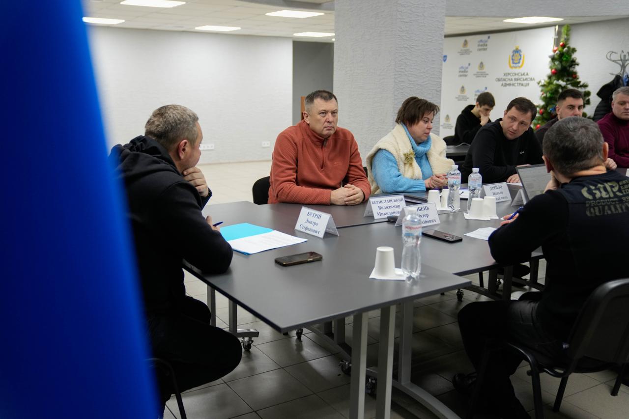Робоча нарада за великим столом у світлому офісному приміщенні. За столом сидять люди, перед ними розміщені таблички з іменами та посадами, папери, смартфони та паперові стаканчики. На задньому плані видно білу стіну з логотипом Херсонської обласної військової адміністрації та прикрашену новорічну ялинку.