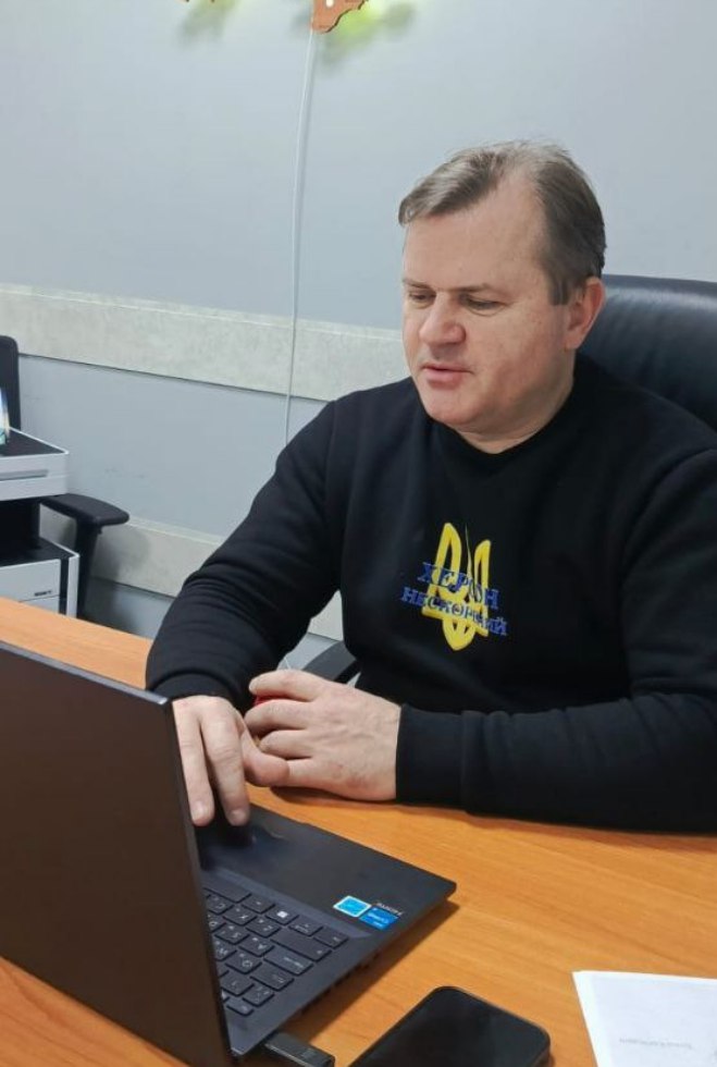 Чоловік у чорному світшоті з гербом України та написом «Херсон незламний» сидить за столом і працює за ноутбуком. Він зосереджено дивиться на екран, поруч на столі лежить смартфон та папери.