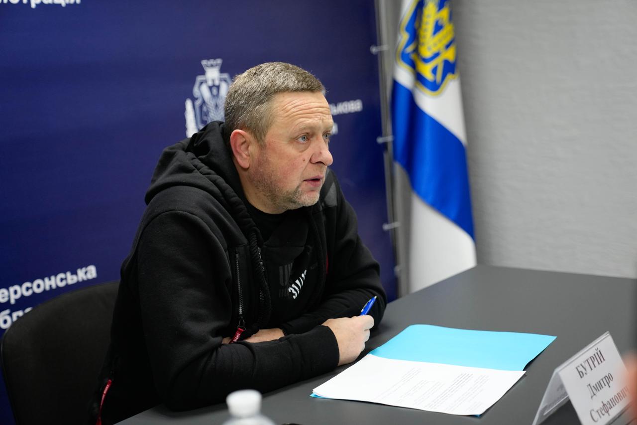 Чоловік у чорній спортивній кофті сидить за столом у офіційному приміщенні. Перед ним лежать документи у блакитній папці та стоїть табличка з іменем. На фоні праворуч розміщено прапор Херсонської області.