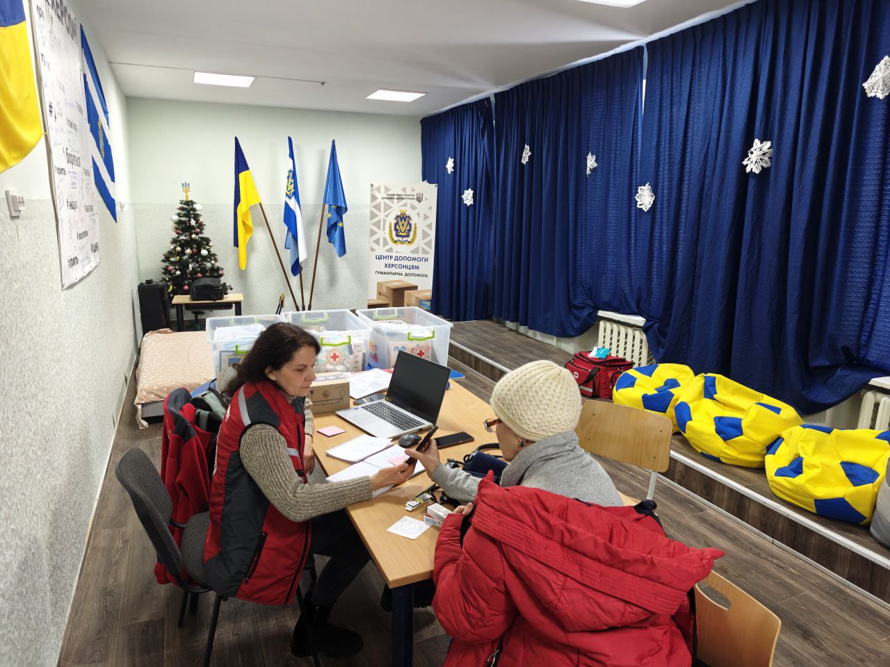 Двоє людей за столом у волонтерському центрі в Херсоні. Жінка у червоному жилеті працює з документами, а навпроти неї сидить інша жінка у червоній зимовій куртці та світлій в'язаній шапці, яка тримає в руці телефон. На столі стоїть ноутбук та лежать папери, а на фоні видно прапори України та ЄС, новорічну ялинку та сині штори зі сніжинками.