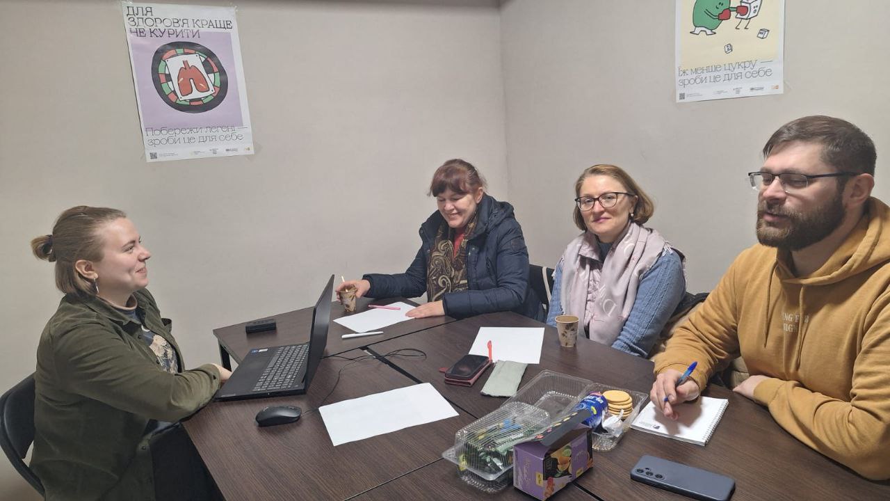 Чотири особи сидять за столом під час робочої зустрічі або обговорення в офісному приміщенні. На столі розміщено відкритий ноутбук, документи, ручки та перекус. На стіні за ними висять інформаційні плакати про здоровий спосіб життя: один про шкоду куріння, інший про зменшення споживання цукру.