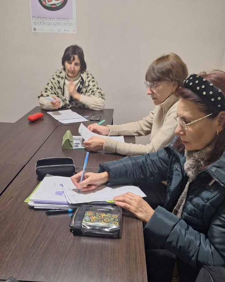 Три жінки сидять у ряд за довгим столом у робочому приміщенні та роблять записи на паперових аркушах. На стіні над ними висить плакат про здоровий спосіб життя із зображенням легень та написом «Побережи легені — зроби це для себе».