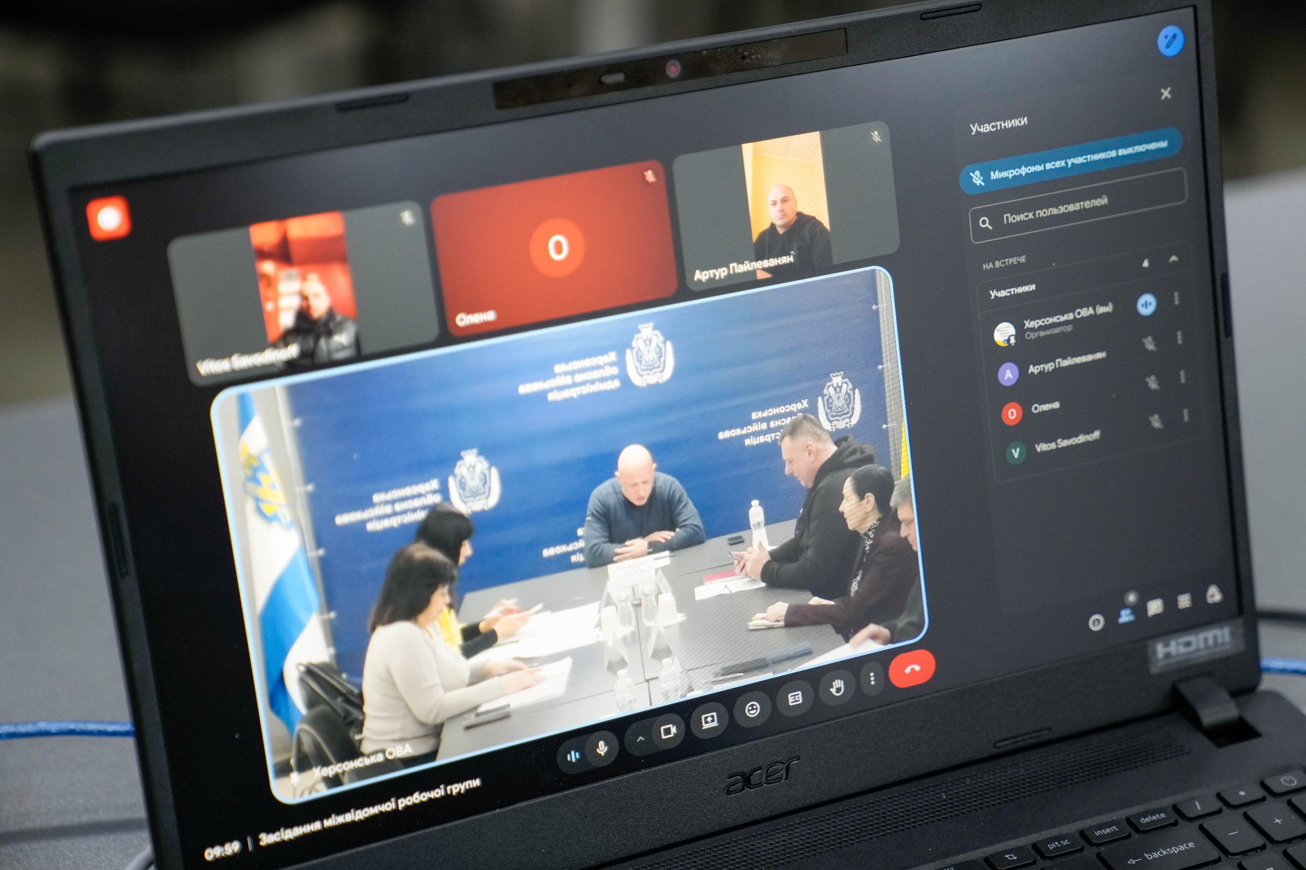 Екран ноутбука, на якому відображено онлайн-конференцію у форматі відеозв'язку. У головному вікні видно групу людей за круглим столом на фоні банера Херсонської ОВА, а зверху — вікна інших дистанційних учасників. У нижній частині екрана напис «Засідання міжвідомчої робочої групи».