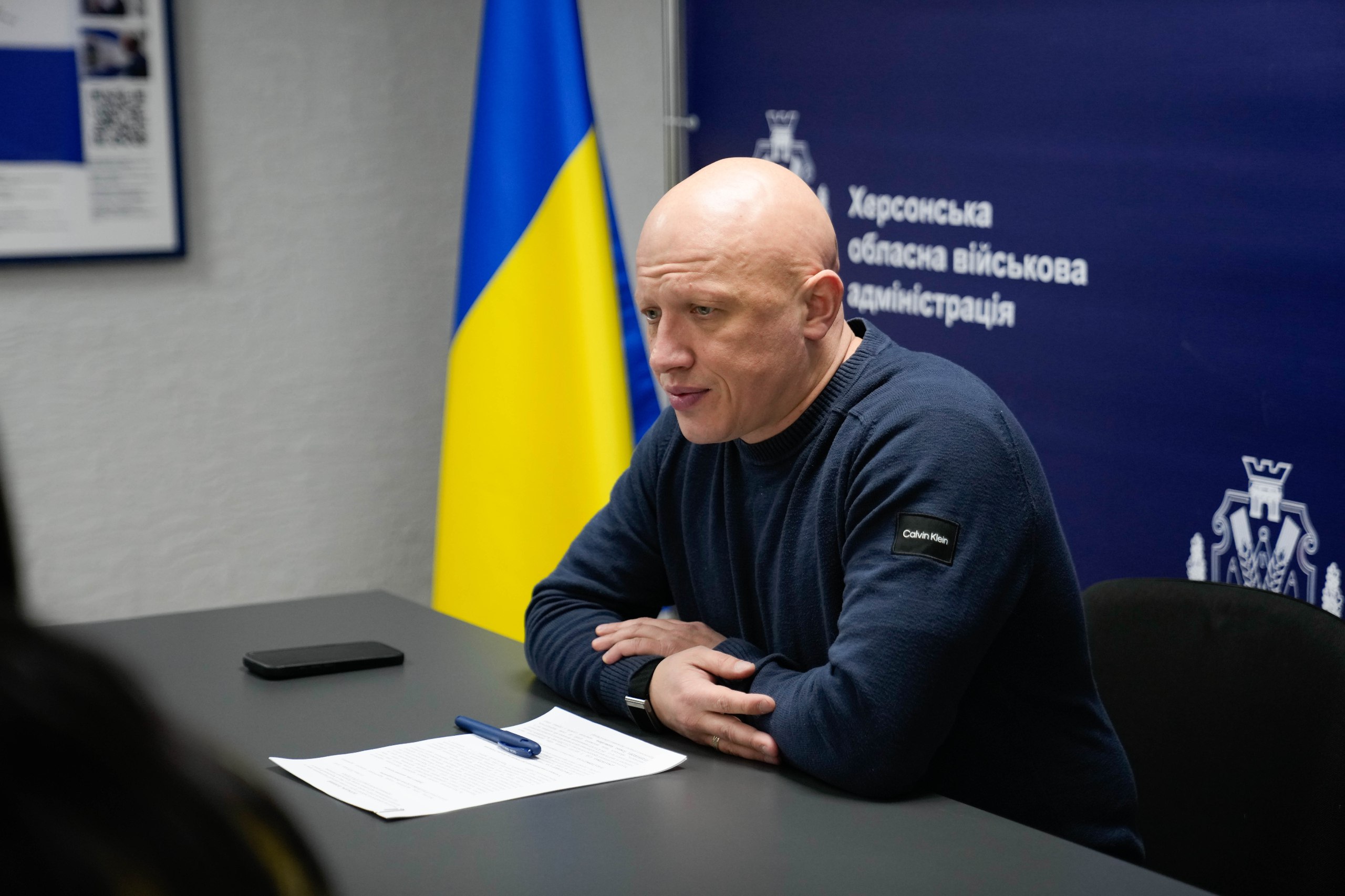 Чоловік у темно-синьому джемпері сидить за столом під час офіційної зустрічі. Перед ним лежить аркуш паперу та ручка. На фоні видно державний прапор України та синій банер із написом «Херсонська обласна військова адміністрація».