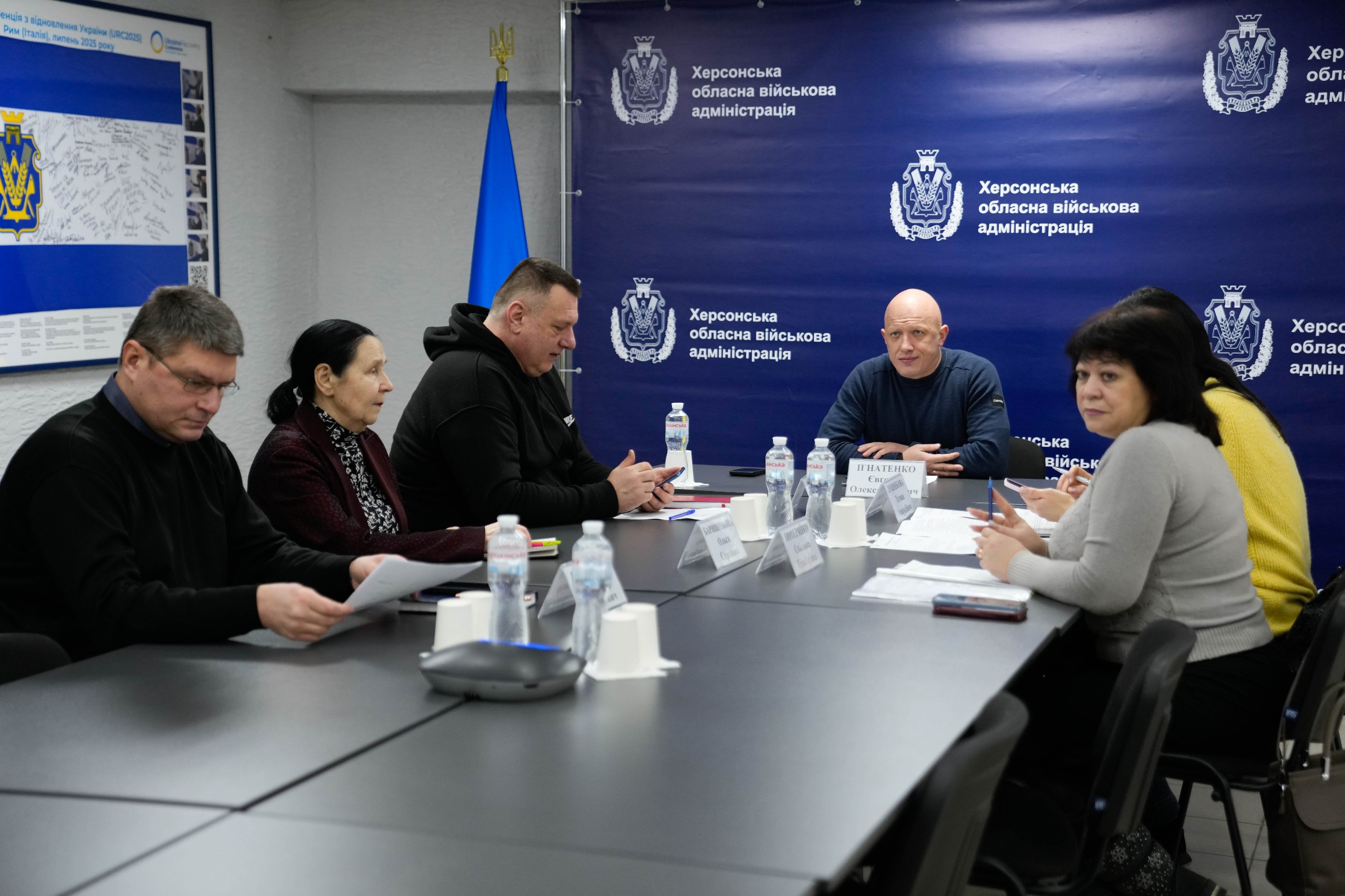 Група людей проводить робочу зустріч за довгим столом у конференц-залі. На фоні розташований синій банер із гербом та офіційним написом «Херсонська обласна військова адміністрація». Учасники переглядають документи та обговорюють питання, на столі стоять пляшки з водою та таблички з іменами.