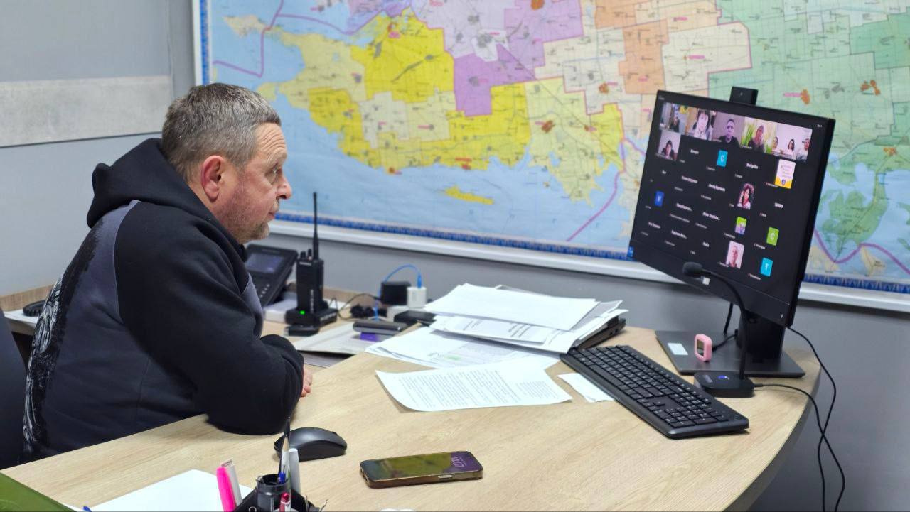 Чоловік у темному худі сидить за робочим столом перед монітором комп'ютера, на якому відображено вікно відеоконференції з багатьма учасниками. На столі лежать документи, клавіатура, миша та рація, а на стіні за ним розміщена велика карта області з різнокольоровими позначеннями.