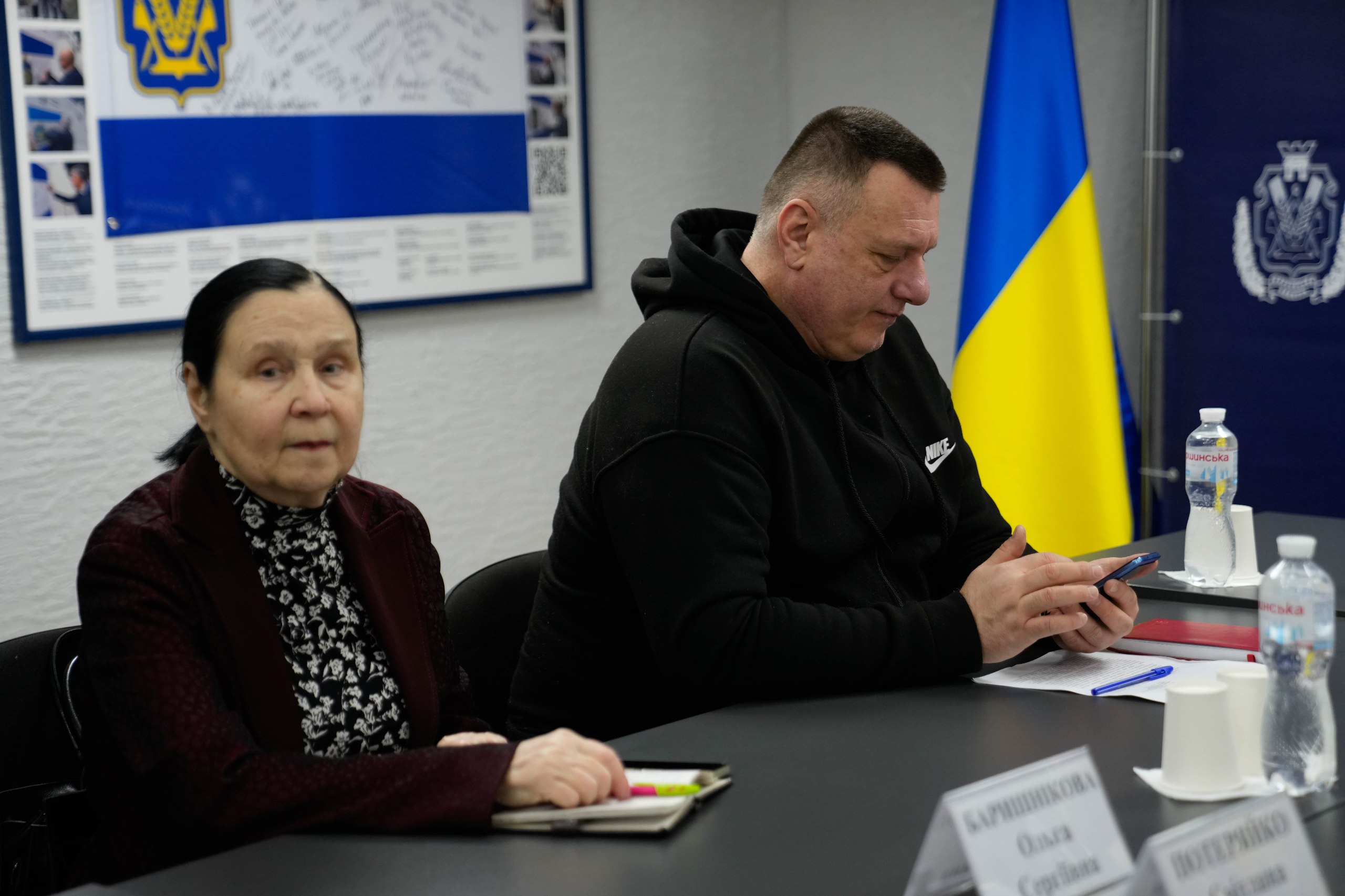 Двоє учасників офіційної зустрічі сидять за столом: жінка в бордовому піджаку з блокнотом та чоловік у чорному худі, який тримає смартфон. Поруч на столі стоять пляшки з водою та інформаційні таблички. На фоні розташований державний прапор України та плакат.
