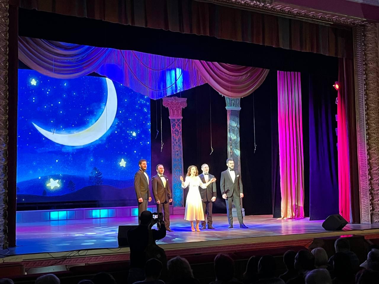 Група артистів — жінка в білій сукні та четверо чоловіків у класичних костюмах — виступають на театральній сцені. На задньому плані розташований великий екран із зображенням нічного неба та півмісяця, а по боках стоять декоративні колони.