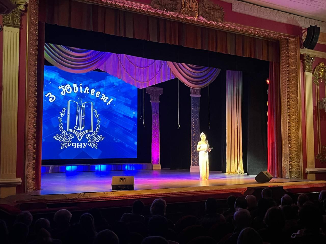 Ведуча у довгій вечірній сукні золотистого кольору стоїть на театральній сцені перед глядацькою залою. На великому екрані позаду неї відображається святкове привітання з ювілеєм та герб університету на синьому фоні.