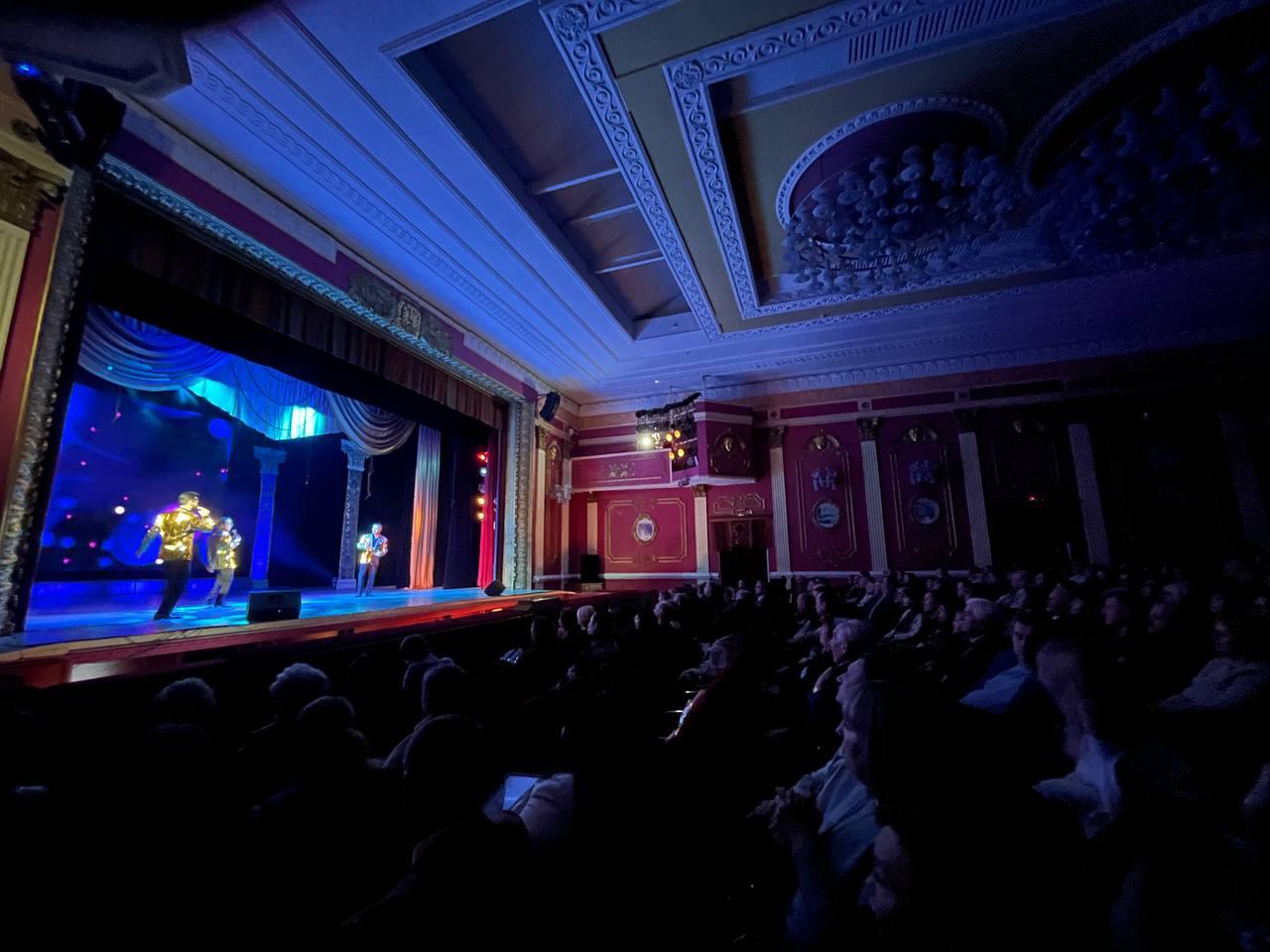 Загальний план глядацької зали театру під час виступу. На освітленій сцені троє артистів у блискучих костюмах виконують номер, а в залі на передньому плані видно силуети глядачів. Інтер'єр приміщення виконаний у класичному стилі з високою декорованою стелею та люстрами.
