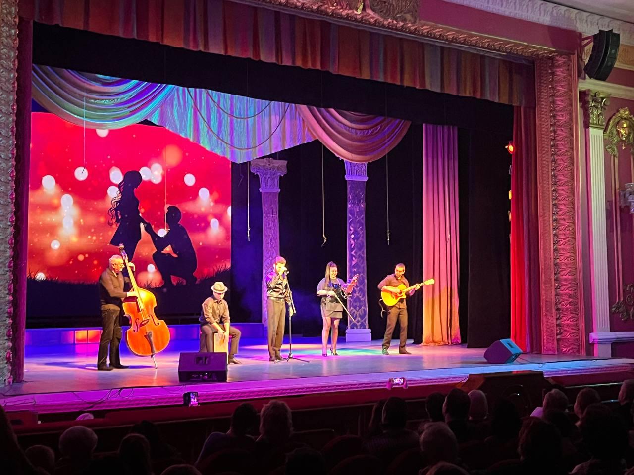 На театральній сцені виступає музичний гурт із п'яти осіб: вокаліст, скрипалька, гітарист, контрабасист та перкусіоніст. На задньому плані розташований великий екран із червоним освітленням та силуетом пари, що заручається.