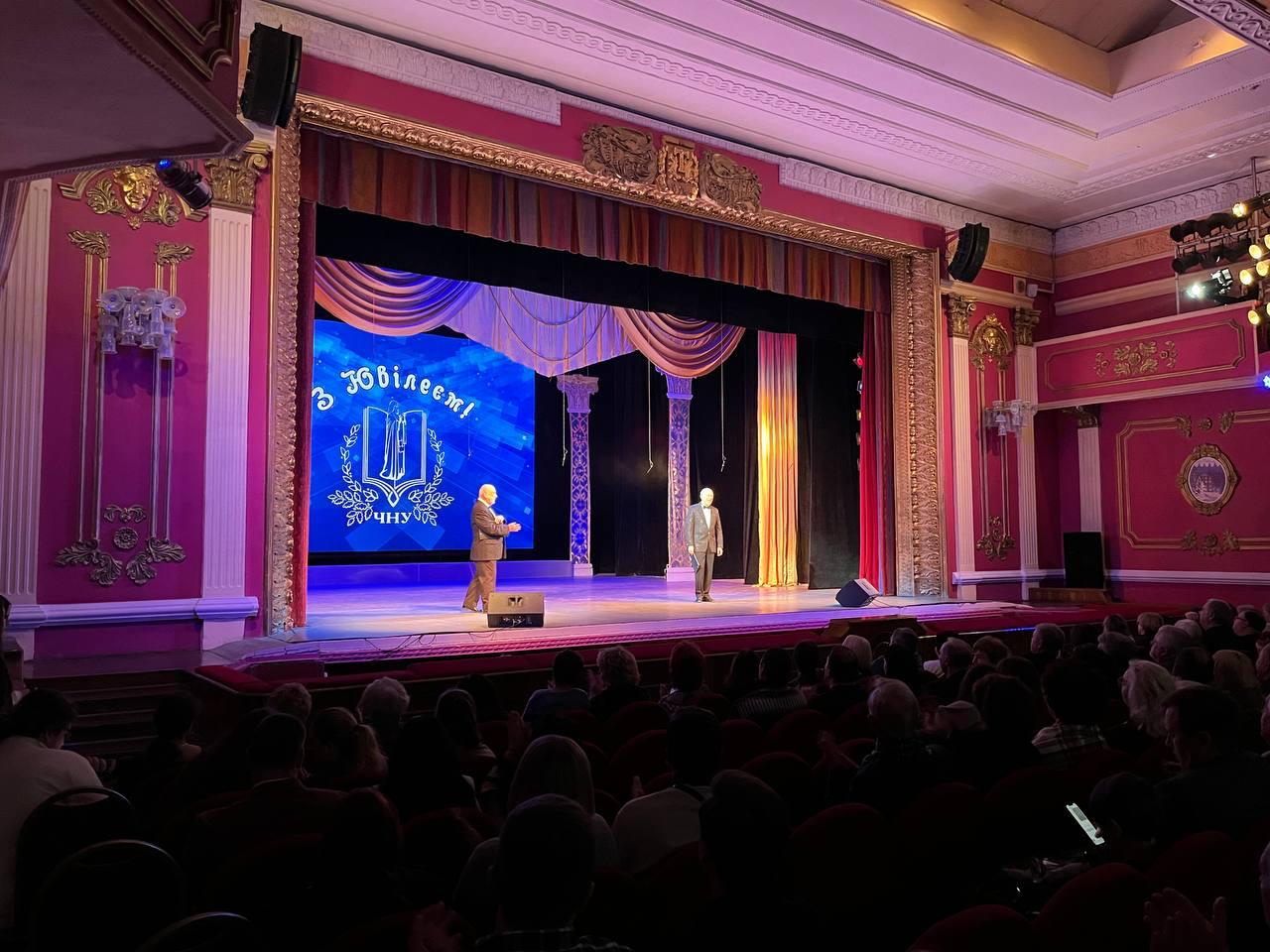 На театральній сцені перед заповненою глядачами залою стоять двоє чоловіків у костюмах. На великому екрані позаду них відображається синє святкове тло з вітальним написом «З ювілеєм!» та академічним гербом.