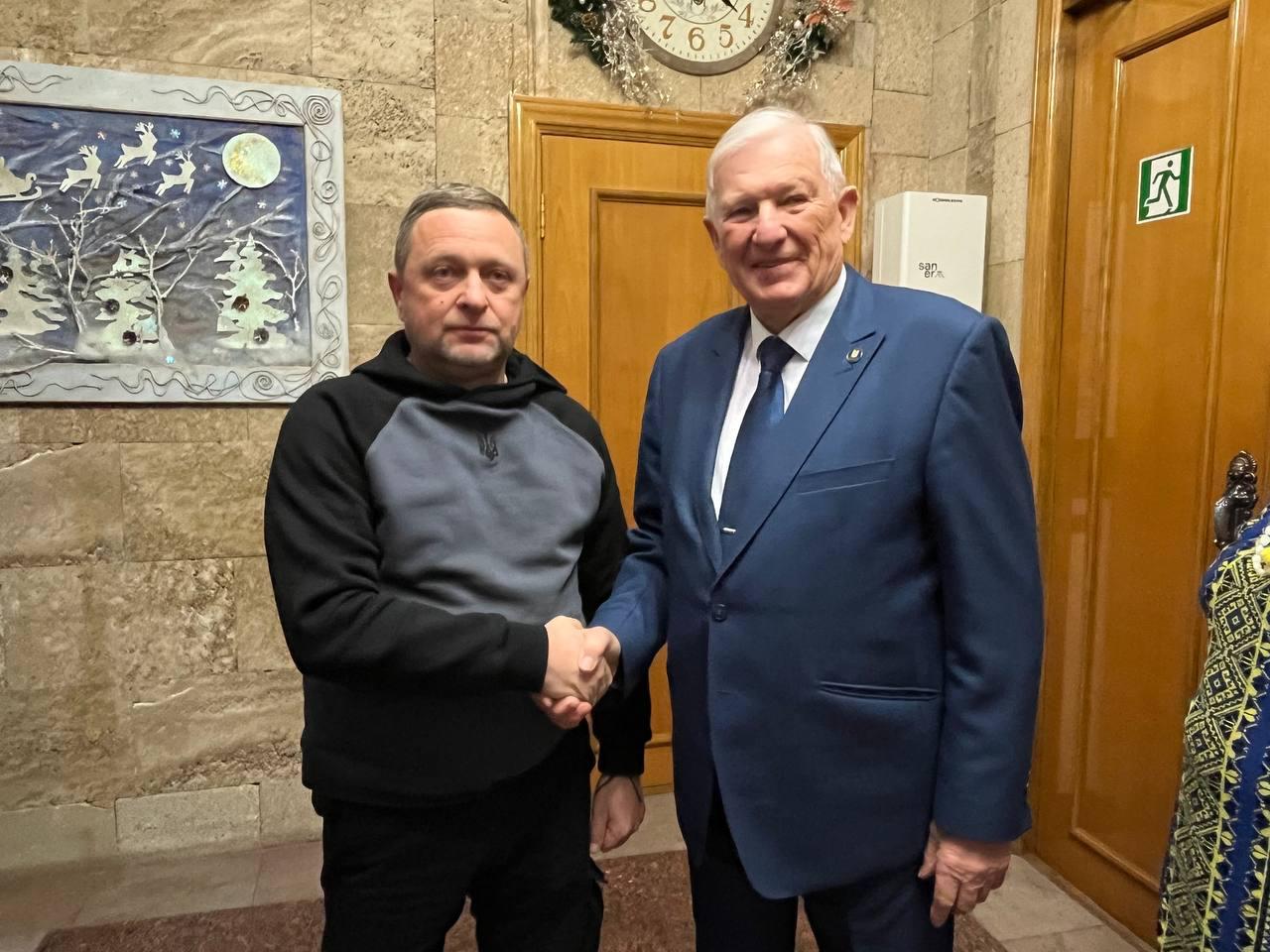 Двоє чоловіків потискають один одному руки в приміщенні. Чоловік ліворуч одягнений у спортивну кофту з капюшоном, а чоловік праворуч — у діловий костюм із краваткою. На стіні позаду них висить картина та годинник.