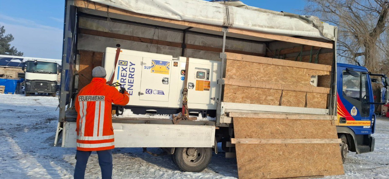 Великий білий промисловий генератор із логотипом «DAREX ENERGY» закріплений ременями у кузові вантажівки. Чоловік у помаранчевому спеціальному одязі стоїть спиною до камери та перевіряє кріплення. Дія відбувається на засніженому майданчику у сонячний день.