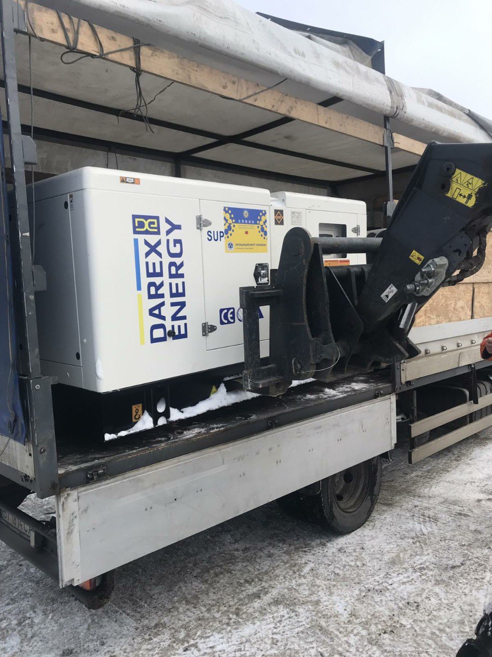 Великий білий промисловий генератор із логотипом «DAREX ENERGY», завантажений у кузов вантажівки. Поруч видно частину навантажувача, що допомагав із транспортуванням. На вулиці зима, на бортах машини та землі лежить сніг.