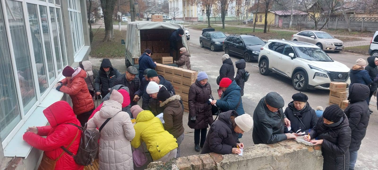 Люди стоять у черзі біля вантажівки, з якої розвантажують коробки з гуманітарною допомогою. Деякі особи заповнюють документи на підвіконнях та огорожі поруч із пунктом видачі.