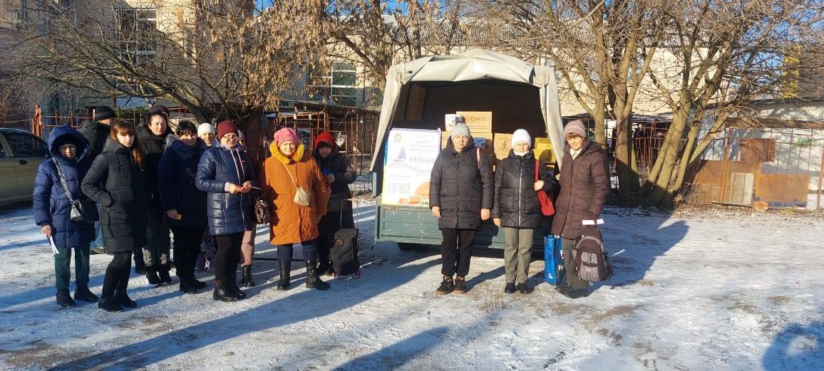 Черга людей стоїть на засніженому майданчику поруч із відкритою вантажівкою, заповненою коробками. На борту автомобіля закріплено плакат «Центр допомоги херсонцям».