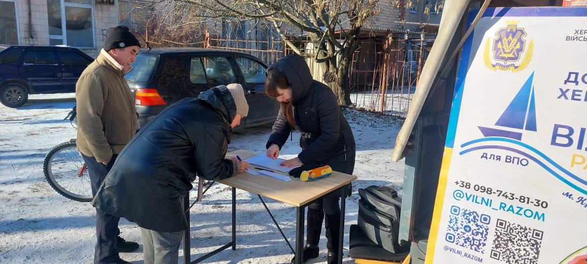 Двоє людей заповнюють бланки на дерев'яному столі на фоні засніженої вулиці та припаркованих авто. Поруч видно частину плаката з контактами волонтерської організації та QR-кодом.