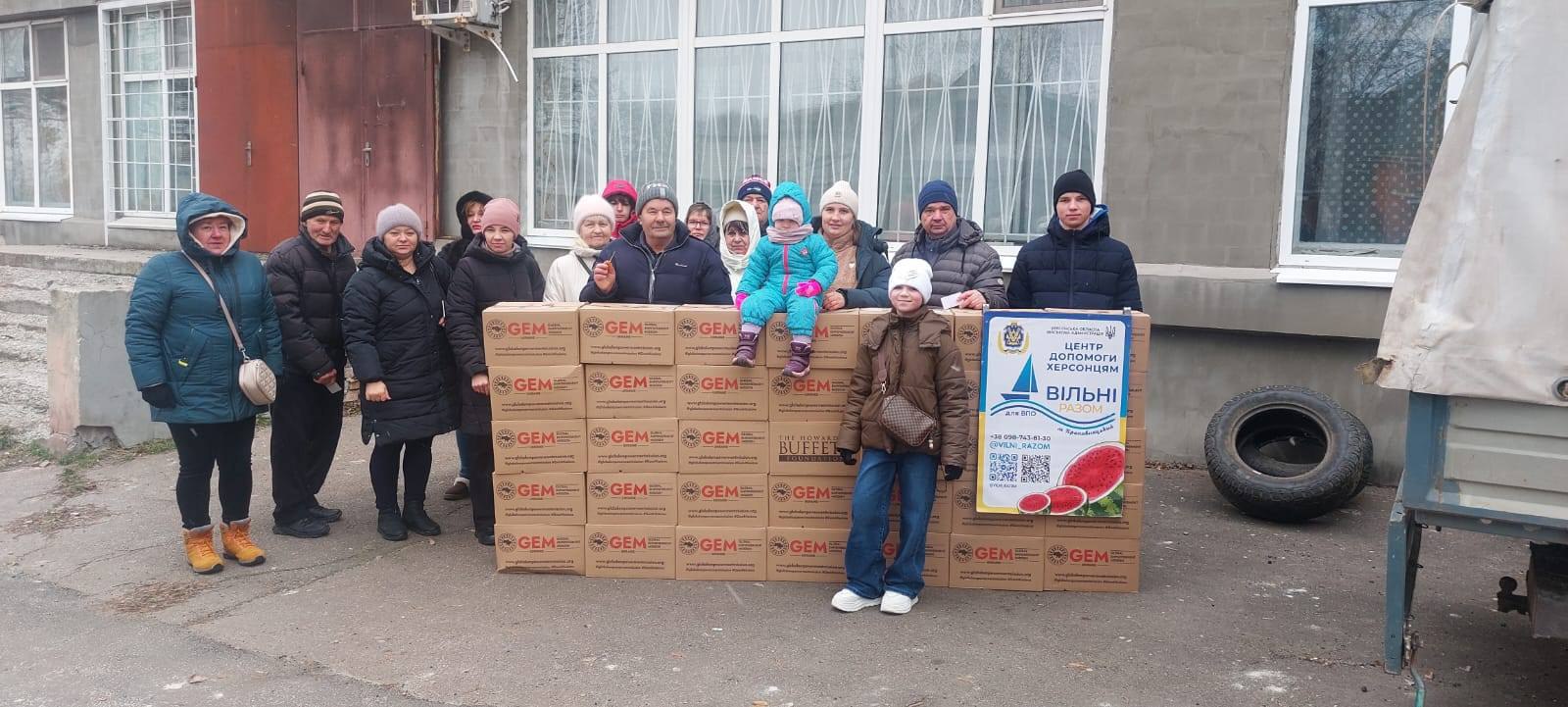 Група людей стоїть поруч із великою кількістю коробок з гуманітарною допомогою від організацій GEM та Howard G. Buffett Foundation. Поруч розміщено інформаційний плакат «Центр допомоги херсонцям».