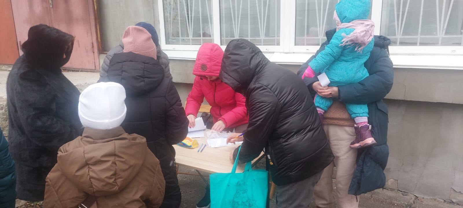 Група дорослих та дітей зібралася біля столу для реєстрації. Люди в зимовому одязі заповнюють документи, необхідні для отримання допомоги.