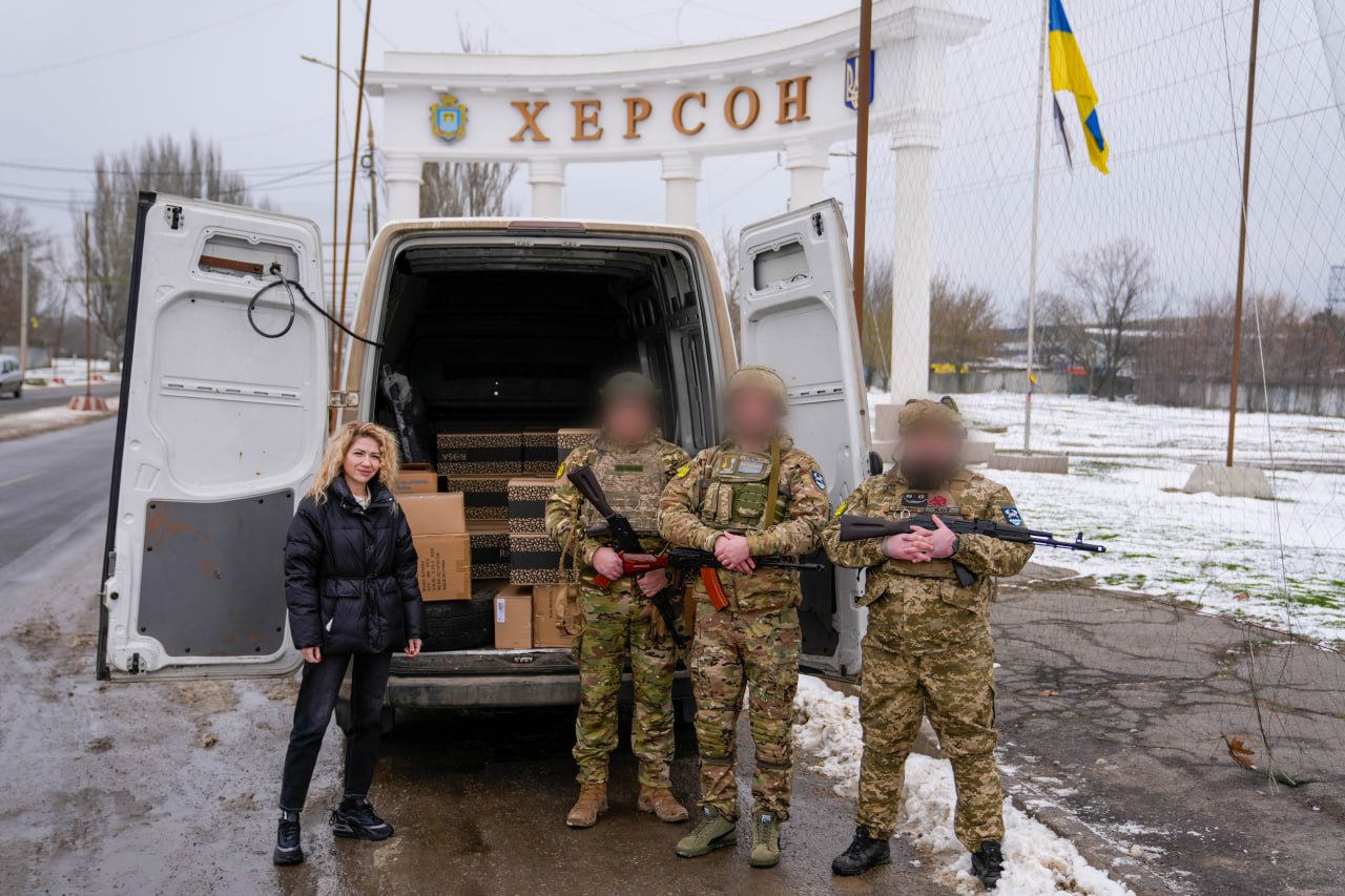 Жінка та троє військових у спорядженні зі зброєю стоять біля відкритого фургона, завантаженого коробками. На задньому плані — в'їзна арка з написом «ХЕРСОН» та державний прапор України. Обличчя військових розмиті з міркувань безпеки.