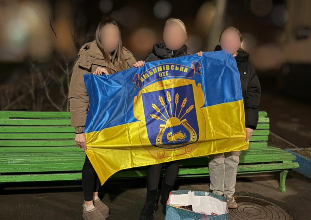 Три жінки в куртках стоять біля зеленої лавки у вечірній час і тримають синьо-жовтий прапор Любимівської ОТГ. У центрі прапора зображено герб громади з колосками пшениці та рибою. Обличчя жінок розмиті для збереження анонімності.