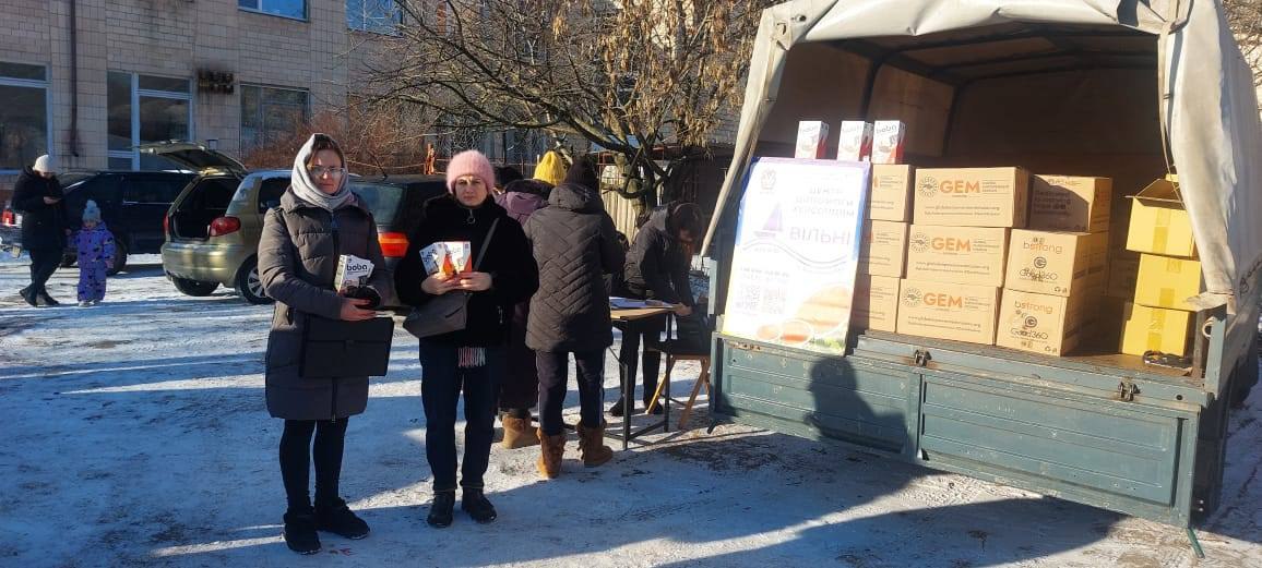 Дві жінки з пакунками в руках стоять біля вантажівки, заповненої коробками з логотипами GEM та Howard G. Buffett Foundation. На задньому плані триває процес реєстрації інших мешканців.