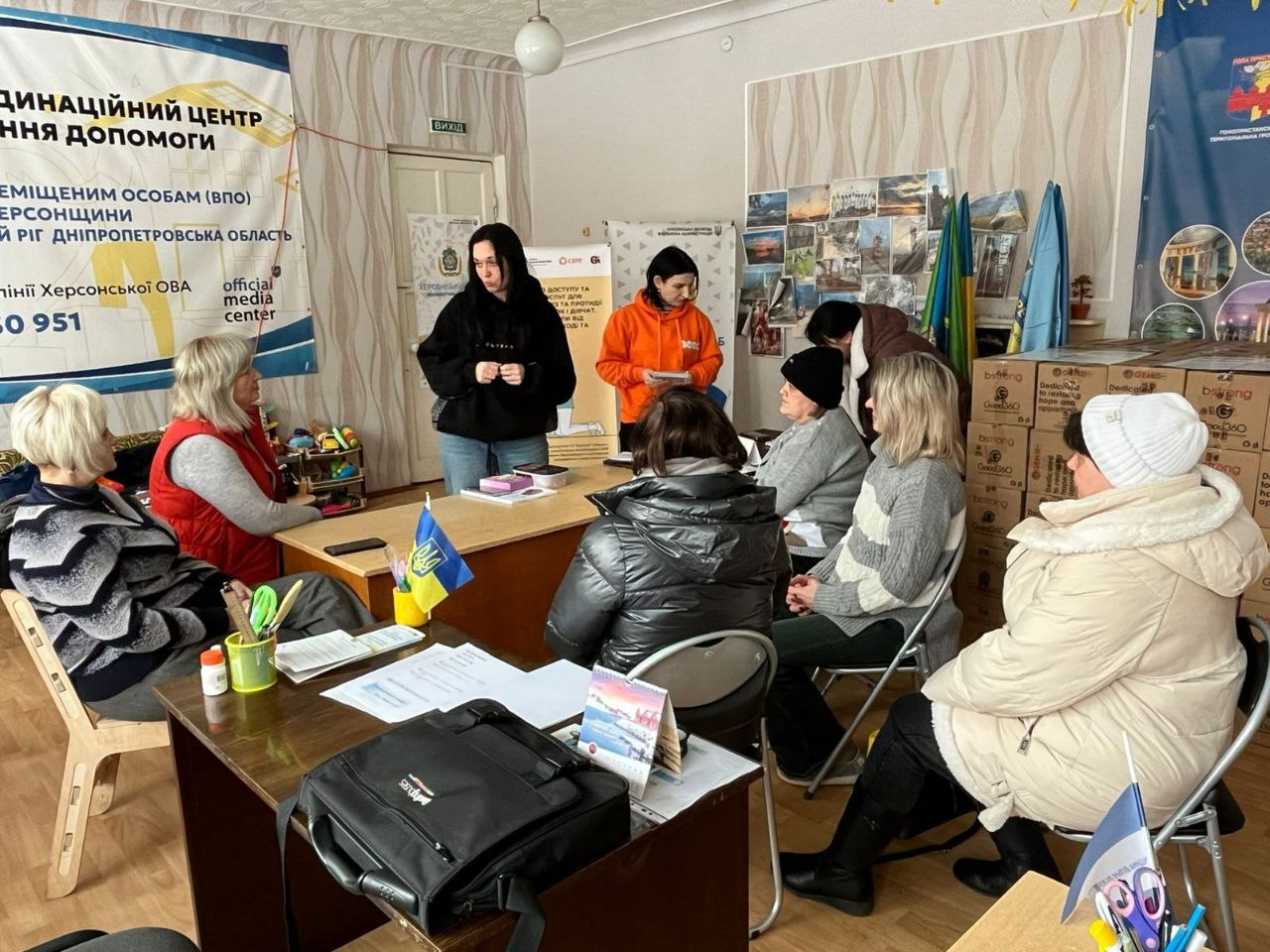 У приміщенні Координаційного центру надання допомоги ВПО з Херсонщини проходить зустріч. Дві фахівчині стоять біля столу та спілкуються з групою жінок, які сидять на стільцях. У кімнаті розміщені інформаційні банери, державна символіка та коробки з гуманітарною допомогою.