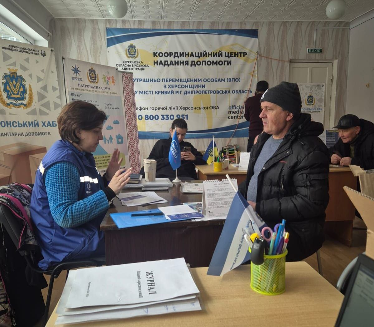 Фахівчиня в синьому жилеті з логотипом міжнародної організації консультує чоловіка в Координаційному центрі допомоги ВПО з Херсонщини у місті Кривий Ріг. На фоні розміщені інформаційні банери з контактами гарячої лінії Херсонської ОВА та логотипами гуманітарних хабів.