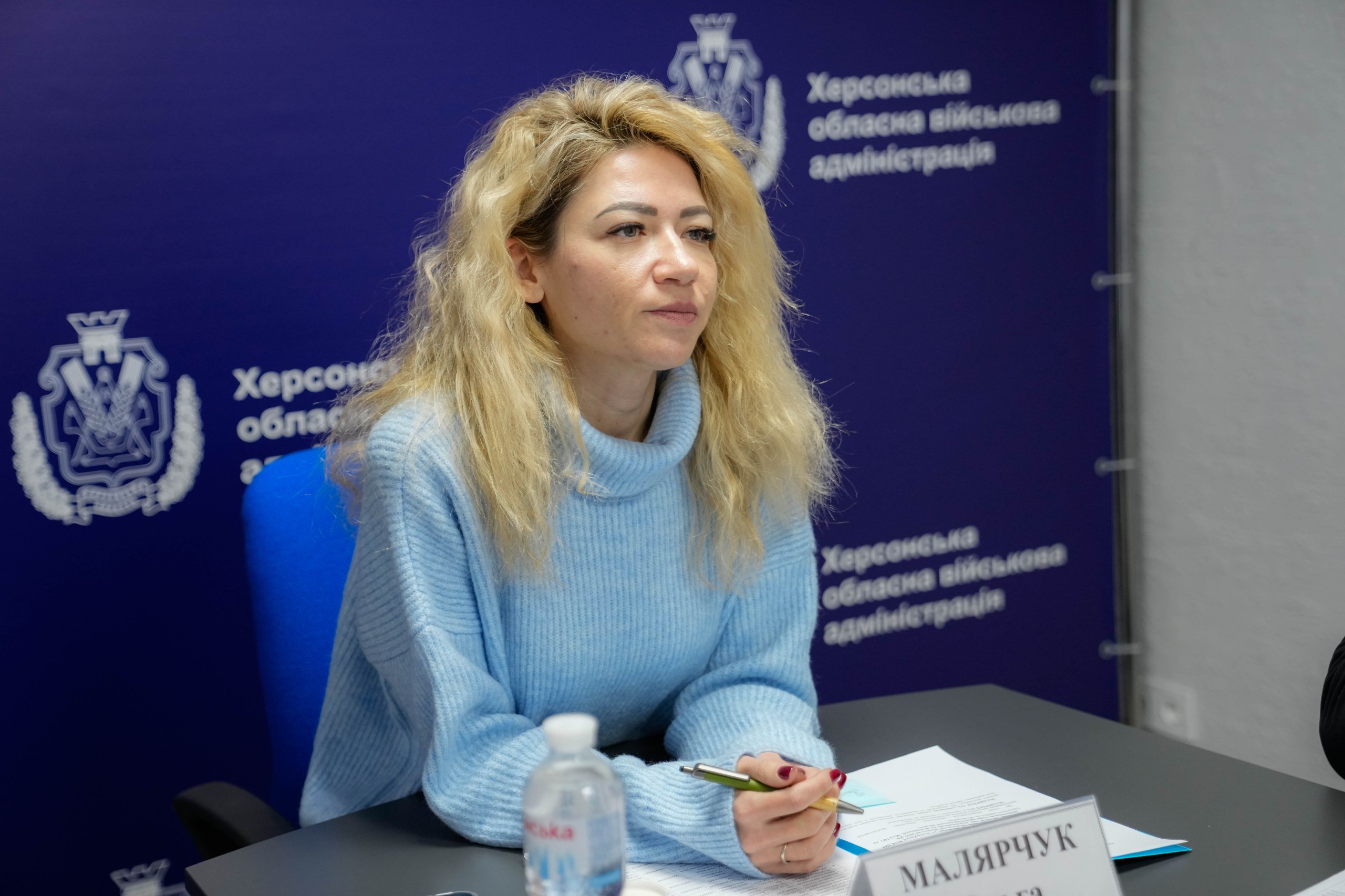 Жінка зі світлим волоссям у блакитному светрі сидить за столом під час робочої наради. Перед нею лежать документи та ручка. На задньому плані — фіолетовий банер із логотипом та написом «Херсонська обласна військова адміністрація».