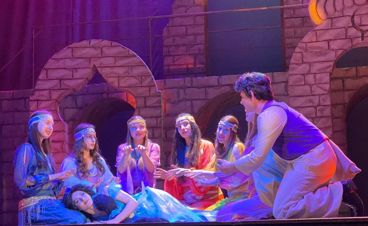 Сцена з театральної вистави за мотивами казки про Аладдіна. Головний герой у білому костюмі та фіолетовій жилетці схилився над принцесою, яка лежить на сцені. Навколо них знаходяться ще п'ятеро дівчат у яскравому східному вбранні. Дія відбувається на тлі декорацій, що зображують кам'яний палац з арками.