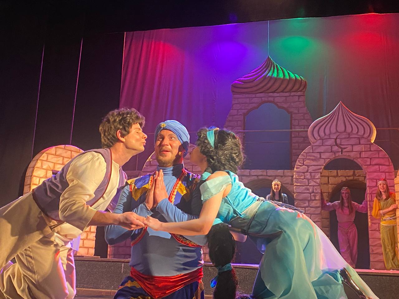 Сцена з театральної вистави за мотивами східної казки. У центрі — актор у костюмі Джина в синьому вбранні та тюрбані, якого з обох боків намагаються поцілувати в щоки інші герої: Аладдін та Жасмін. На задньому плані декорації у вигляді східного палацу з куполами та інші актори в яскравих костюмах.
