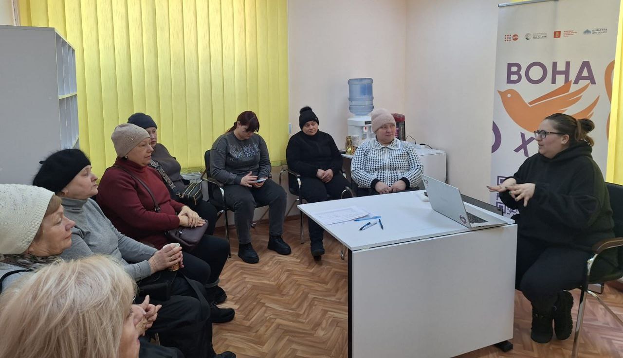 Жінка проводить презентацію для групи слухачок, сидячи за столом із ноутбуком. У приміщенні встановлено банер із написом «ВОНА», а учасниці в теплому одязі уважно слухають доповідачку.